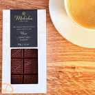 Moksha Chocolate 80% Single Origin Cacao Dark Chocolate from Camino Verde, Ecuador, in elegant white and black packaging, displayed on a wooden surface next to a cup of coffee.