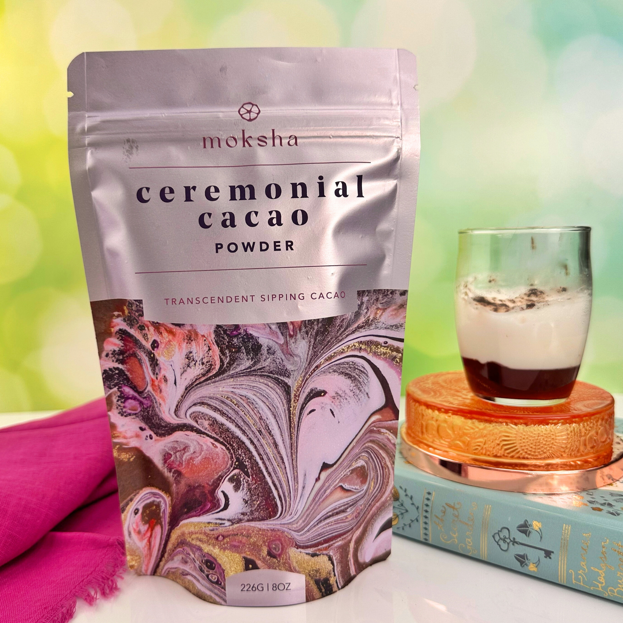 Package of Moksha Ceremonial Cacao Powder (226g/8oz) with vibrant marbled art. A glass of cacao sits beside it on a colorful background—perfect for ceremonial cacao rituals.